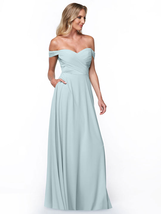 A woman in Avery Austin’s sky blue 83018 gown with off-the-shoulder, fitted pleated bodice, A-line skirt, and pockets stands against a white background, her hand tucked into the dress pocket as the floor-length gown flows elegantly.