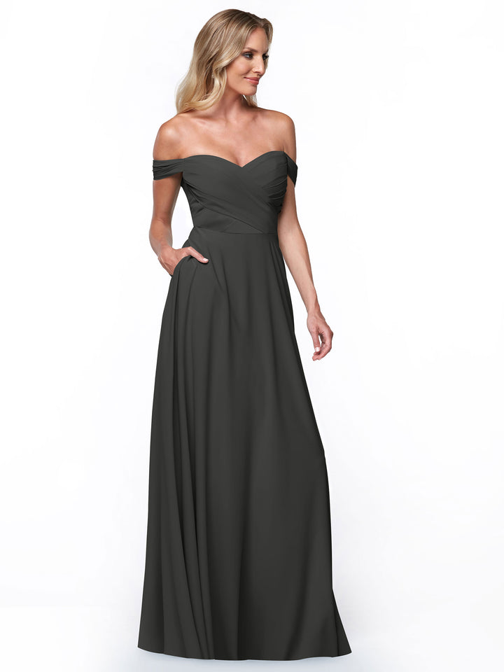 A woman models the Avery Austin 83018 dress, featuring an off-the-shoulder neckline, fitted bodice, A-line skirt, and pockets, as she stands against a plain white background, gazing thoughtfully to the side.