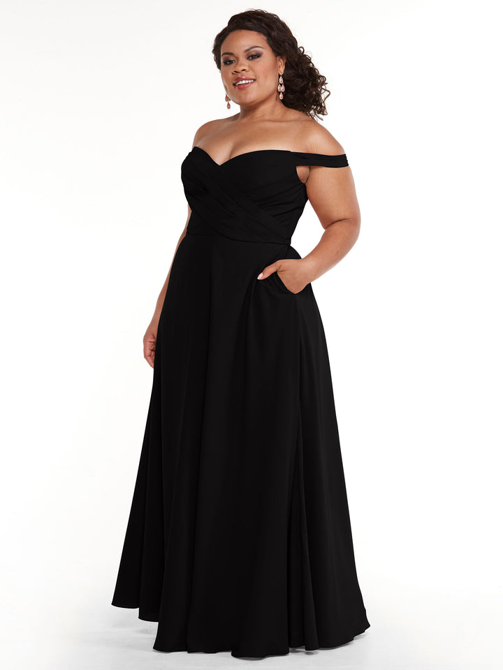 A woman with curly hair smiles in Avery Austin style 83018, an elegant off-the-shoulder dress featuring a fitted bodice, A-line skirt, and pockets. She wears drop earrings against a plain white background.