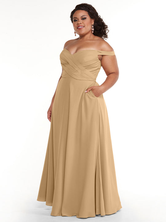 A woman wears Avery Austin’s floor-length dress (style 83018) with off-the-shoulder design, fitted ruched bodice, A-line skirt, and pockets. She has one hand in a pocket and accessorizes with earrings.