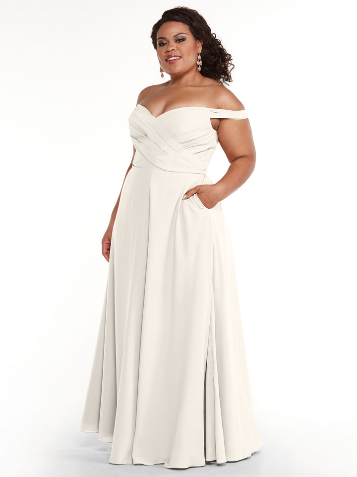 A woman smiles in the Avery Austin 83018 gown, an ivory off-the-shoulder dress with a fitted bodice, pleated details, A-line skirt, and convenient pockets, posing gracefully with one hand tucked in her pocket.