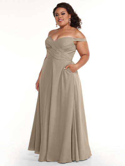 A woman models Avery Austin's 83018 dress, featuring a taupe off-the-shoulder design with a pleated bodice, A-line skirt, and side pockets, posed against a white background.