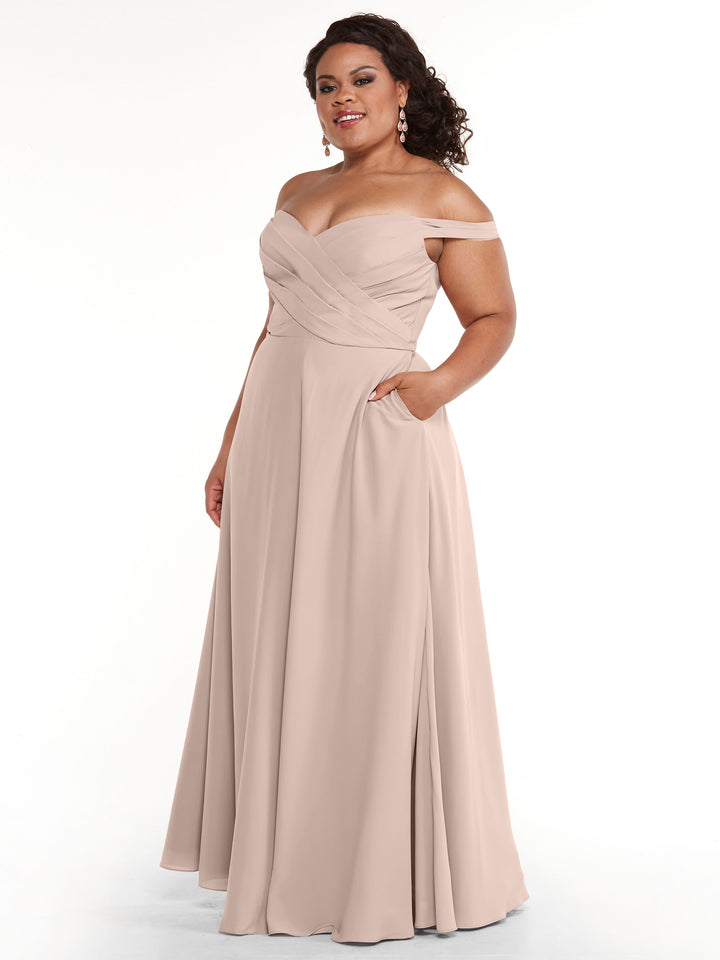 A woman models the Avery Austin 83018 dress in rose pink, featuring an off-the-shoulder style, fitted pleated bodice, A-line skirt, and pockets, posing confidently with a plain white backdrop.