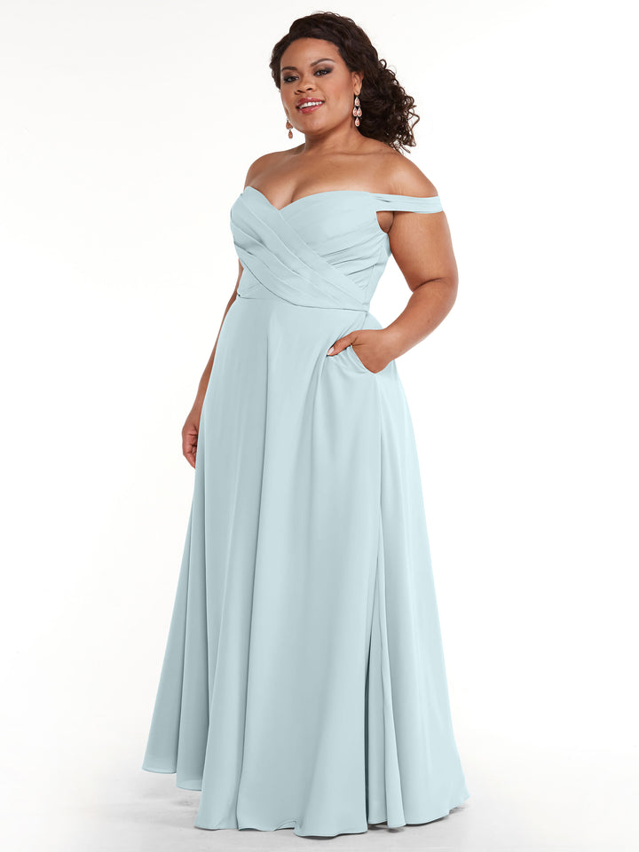 A woman models Avery Austin’s 83018: a light blue, off-the-shoulder A-line gown with a fitted bodice, pleated details, side pockets, and a floor-length skirt, standing gracefully against a white background.
