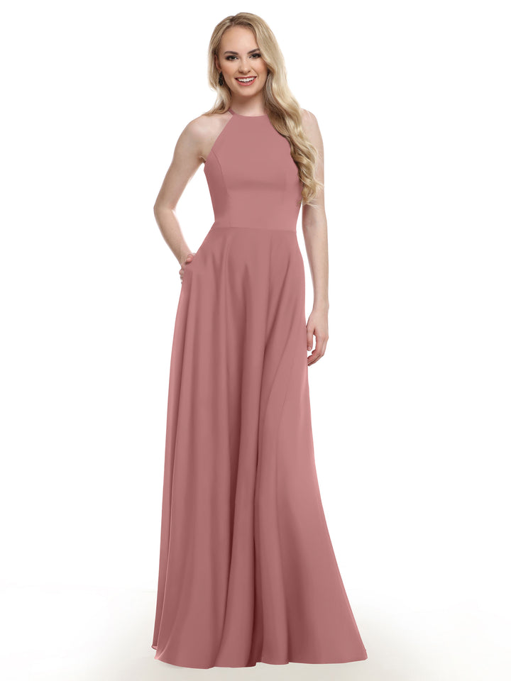 A woman with long blonde hair is smiling and posing with one hand on her hip, wearing Avery Austin’s sleeveless, floor-length dusty rose pink A-line gown (Style 83022) against a white background.