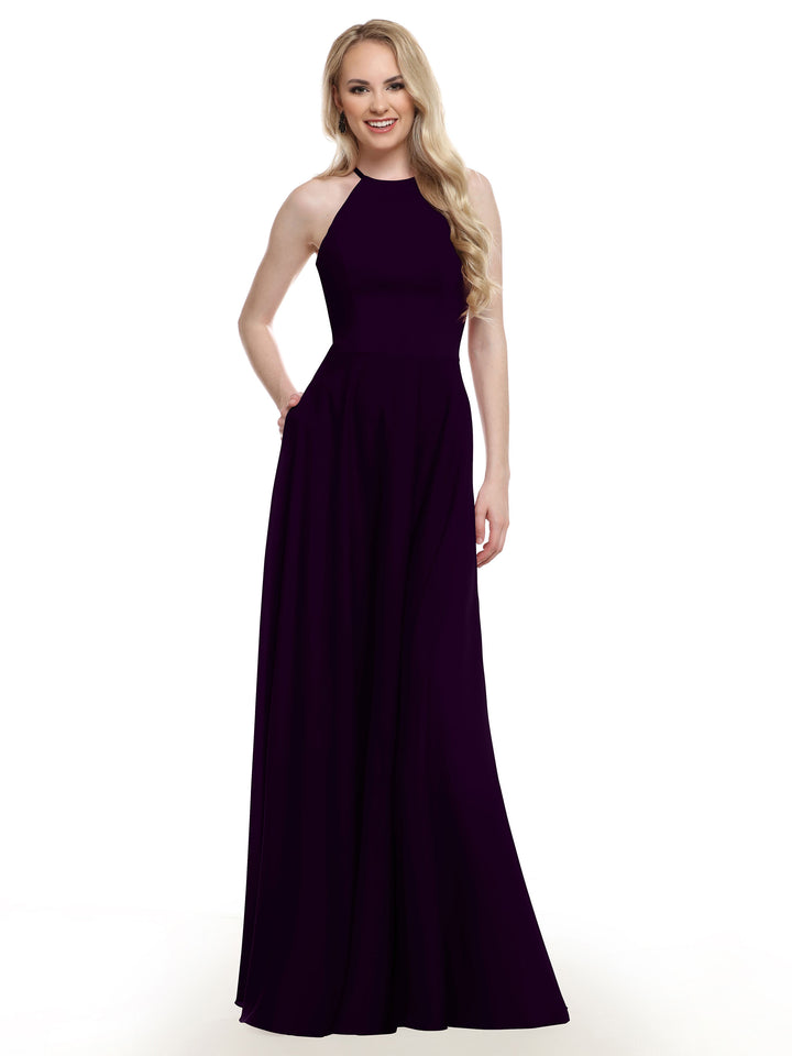 A woman stands smiling with one hand in her pocket, wearing Avery Austin style 83022—a sleeveless, floor-length dark purple A-line gown—against a plain white background.
