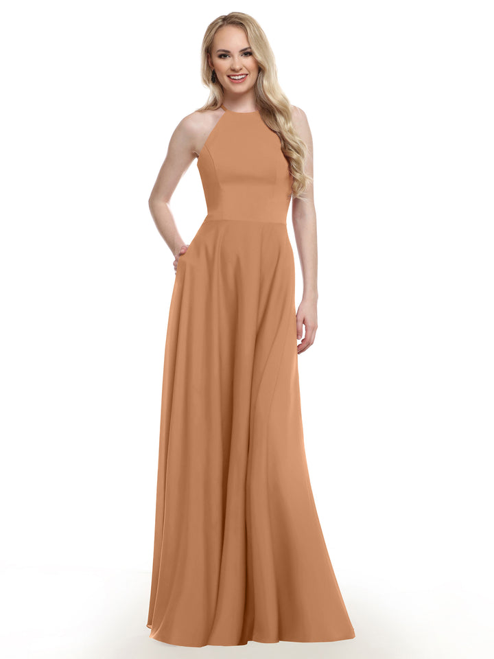 A woman with long blonde hair smiles while wearing Avery Austin’s 83022 sleeveless, floor-length peach A-line gown with a high neckline, posing with one hand in her pocket against a plain white background.