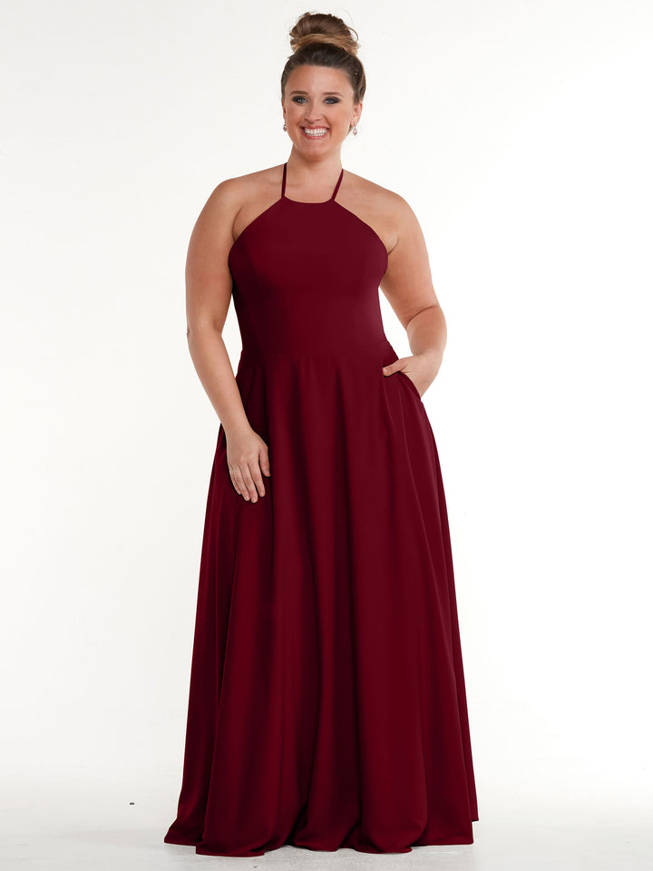 A smiling woman with a high bun wears Avery Austin style 83022, a sleeveless deep red halter dress with an A-line skirt and pockets, standing against a plain white background.