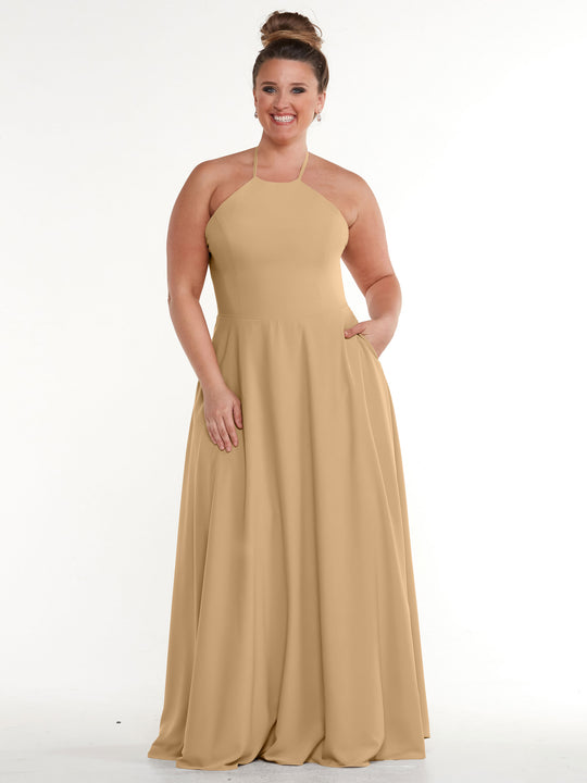 A woman with her hair in a bun smiles in the Avery Austin TH-83022: a sleeveless, floor-length beige halter dress with an A-line skirt and pockets, standing against a plain white background.