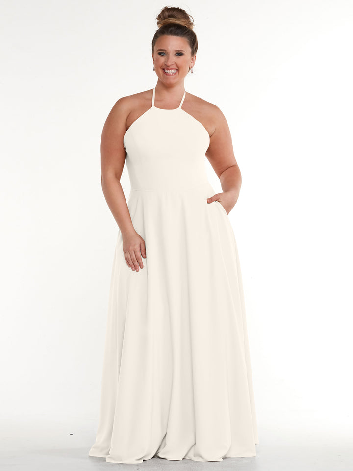 A woman smiles with one hand in her pocket while wearing the Avery Austin 83022—a sleeveless, floor-length white dress with an A-line skirt and halter neckline—standing against a plain white background.