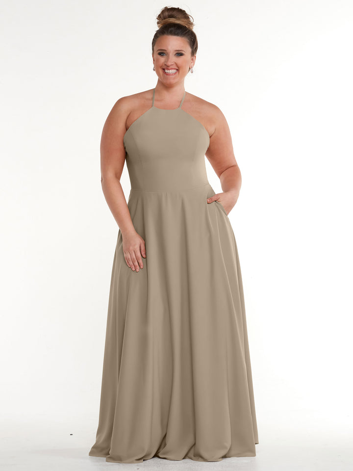 A woman smiles with one hand in her pocket, wearing the Avery Austin 83022: a taupe halter-neck, floor-length dress with an A-line skirt, posed against a plain white background.