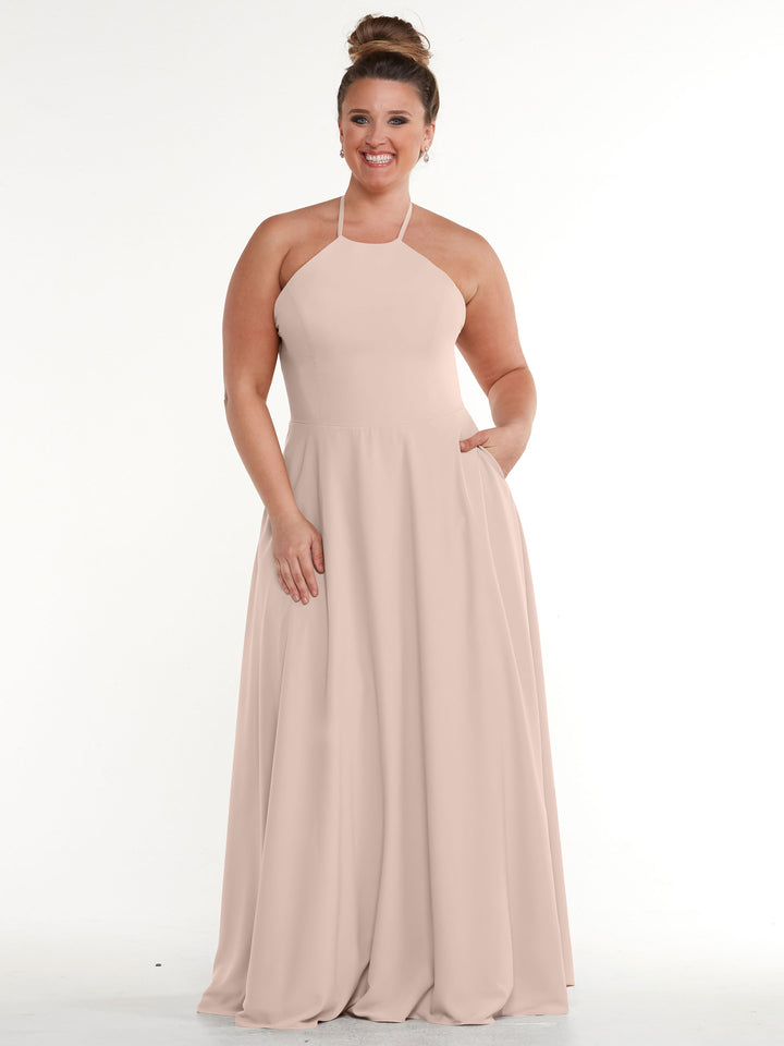A woman with her hair in a bun smiles in a sleeveless Avery Austin 83022 rose pink halter dress with an A-line skirt and pockets, standing against a plain white background.