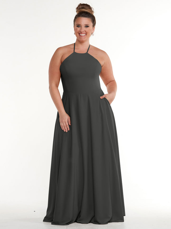 A woman with her hair in a bun wears the Avery Austin 83022, a sleeveless, floor-length dark gray dress with a halter neckline and A-line skirt with pockets, smiling at the camera against a plain white background.