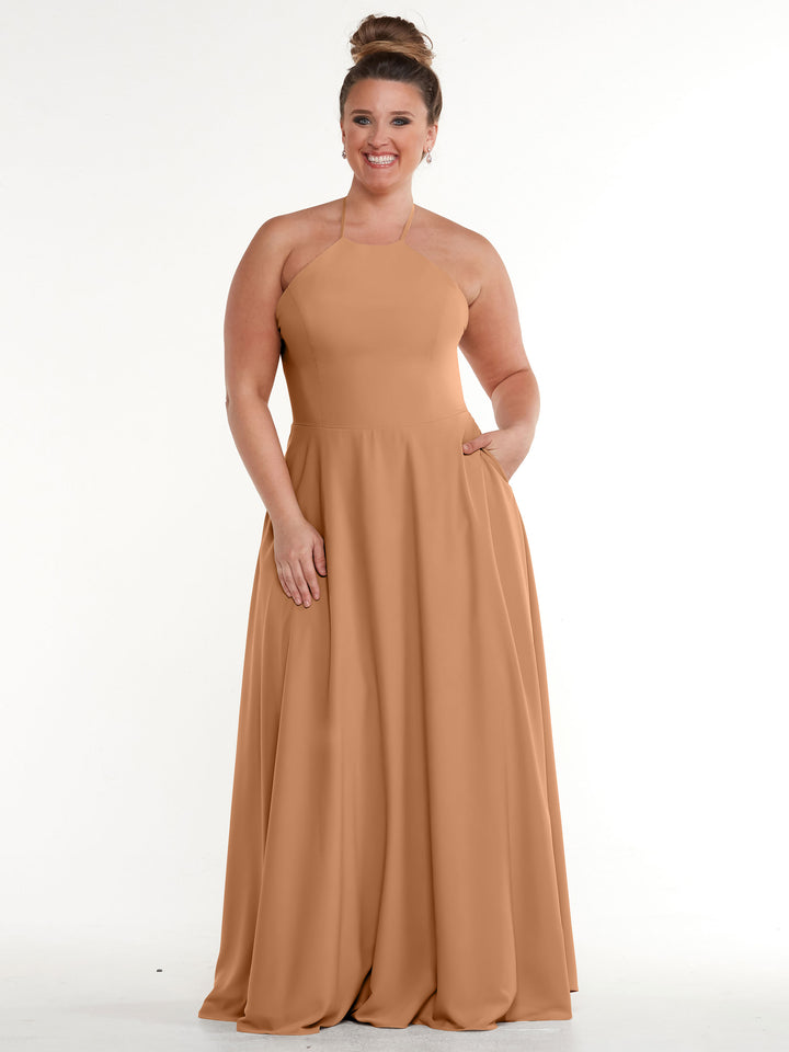 A woman with light skin and brown hair in a bun smiles while wearing the Avery Austin 83022, a sleeveless, floor-length tan halter dress with an A-line skirt and pockets, standing against a plain white background.
