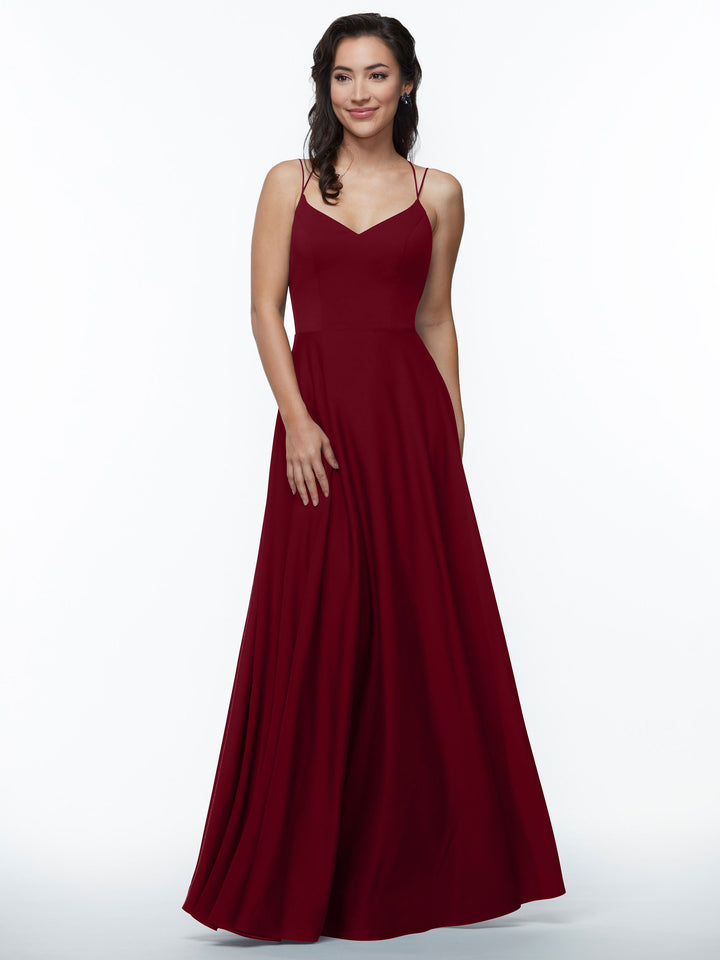 A woman wears Avery Austin style 83024, an elegant floor-length deep red A-line dress with double spaghetti straps, standing against a plain white background and smiling gently with one hand on her gown.