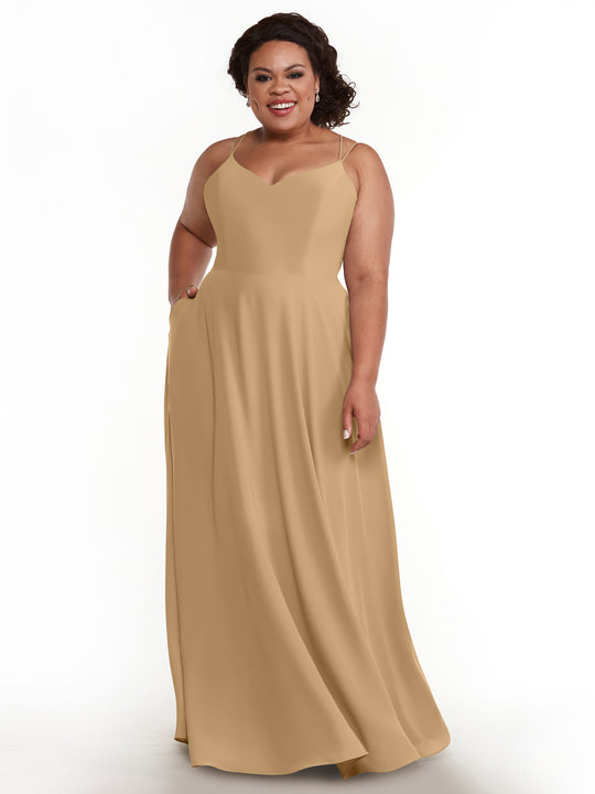 A smiling woman with dark curly hair wears the Avery Austin 83024, an elegant light tan, sleeveless A-line gown with double spaghetti straps, standing against a plain white background.