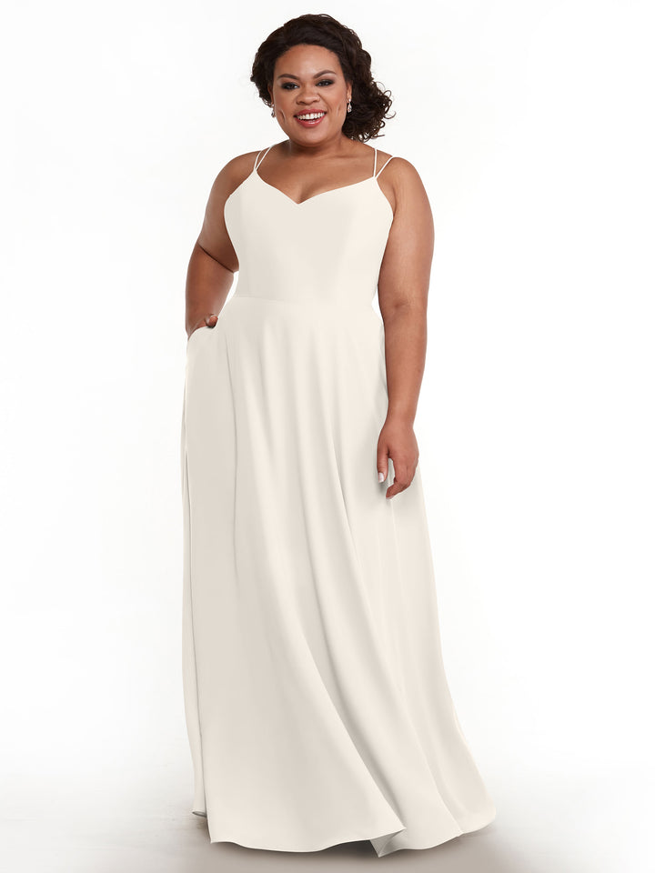 A woman with curly dark hair smiles while wearing Avery Austin’s floor-length A-line dress (style 83024) with double spaghetti straps, a fitted bodice, and a flowing skirt, posed against a plain white background.