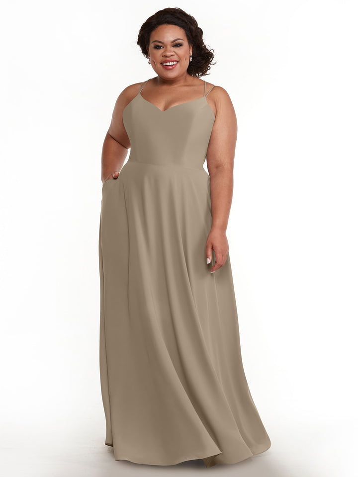 A woman with curly dark hair smiles while wearing Avery Austin’s elegant, floor-length A-Line dress (Style 83024) featuring double spaghetti straps and a flowing skirt. She stands against a plain white background.