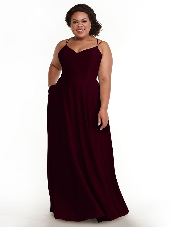 A woman with curly hair smiles in the Avery Austin 83024, an elegant sleeveless floor-length burgundy A-line dress with double spaghetti straps, standing against a plain white background.