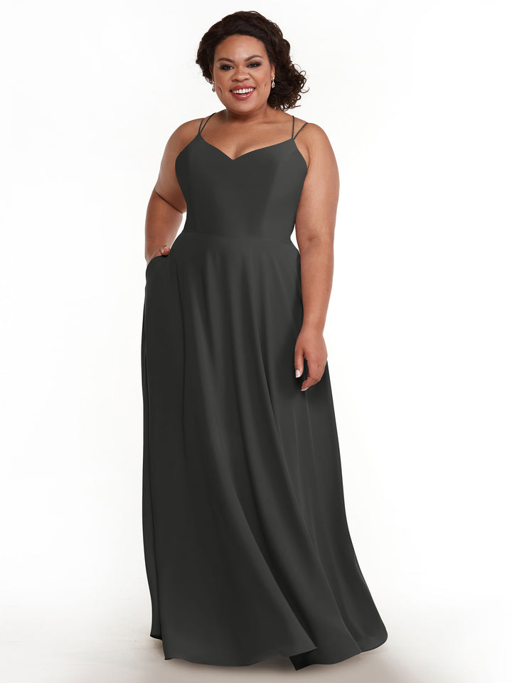 A woman with curly dark hair smiles against a white background, left hand in her pocket, wearing Avery Austin’s elegant A-Line dress, style 83024, featuring double spaghetti straps.