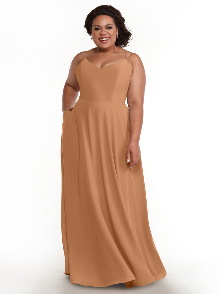 A woman with curly dark hair smiles while modeling the Avery Austin 83024, an elegant floor-length A-line dress with a fitted bodice, double spaghetti straps, and a flowing skirt, against a plain white background.