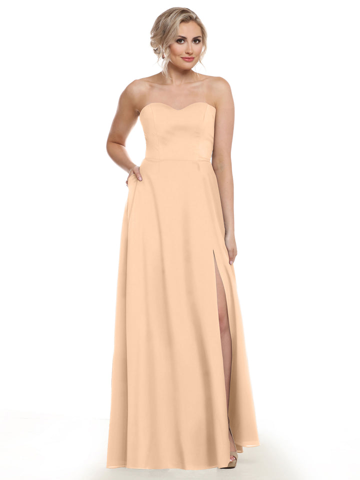 A woman models Avery Austin style 83025, a strapless chiffon bridesmaid dress in light peach with a sweetheart neckline, A-line silhouette, and high side slit, posed against a white background.