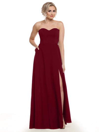 A woman models Avery Austin 83025, a floor-length burgundy chiffon bridesmaid dress with a strapless sweetheart neckline and high side slit. She poses against a white background, her blonde hair styled in loose waves, hand in pocket.