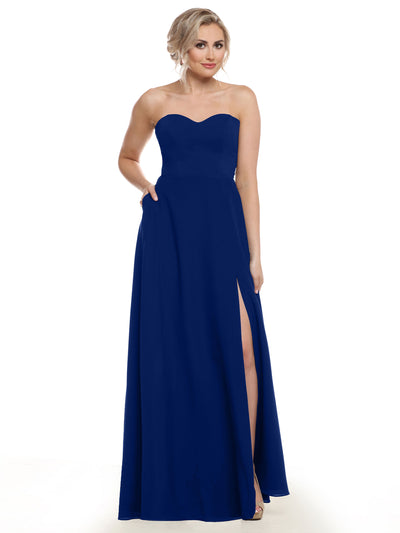 A woman models Avery Austin’s floor-length royal blue bridesmaid dress (style 83025), featuring a strapless, sweetheart neckline and high slit; she poses with one hand in her pocket, the other holding the chiffon skirt.