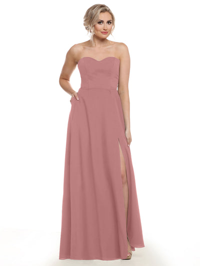 A woman wears Avery Austin style 83025, a mauve pink strapless bridesmaid gown with a sweetheart neckline, thigh-high slit, floor-length skirt, and pockets. She stands confidently, blonde hair in an updo.