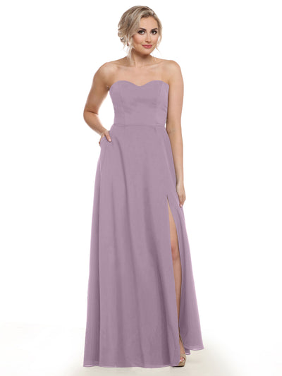 A woman in Avery Austin’s 83025—a strapless, lavender, floor-length bridesmaid dress with a sweetheart neckline, thigh-high slit, and pockets—stands against a white background with one hand in her pocket.