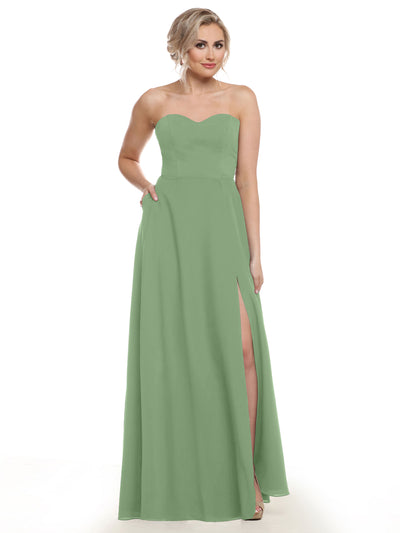 A woman stands against a white background in Avery Austin's 83025—a strapless, sage green chiffon bridesmaid dress with a sweetheart neckline, thigh-high slit, floor-length hem, and pockets, one hand in her pocket and the other by her side.