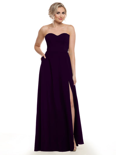 A woman models Avery Austin's style 83025—a strapless, floor-length dark purple chiffon bridesmaid dress with a sweetheart neckline and thigh-high slit. She stands against a white background with short blonde hair and one hand in her pocket.