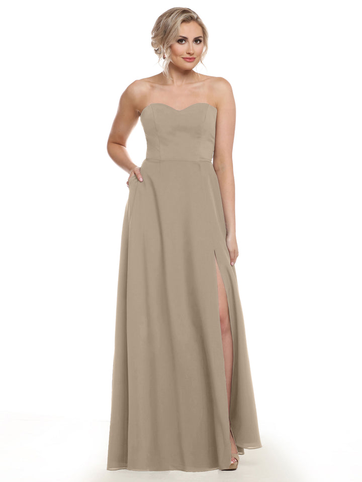 A woman poses in Avery Austin’s 83025, a strapless, floor-length taupe bridesmaid dress featuring a sweetheart neckline, high side slit, and pockets, set against a white background.