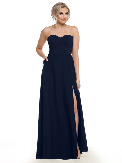 A woman poses against a white background in Avery Austin’s 83025: a strapless, floor-length navy chiffon bridesmaid dress with a sweetheart neckline and thigh-high slit, one hand in her pocket and the other holding the dress.