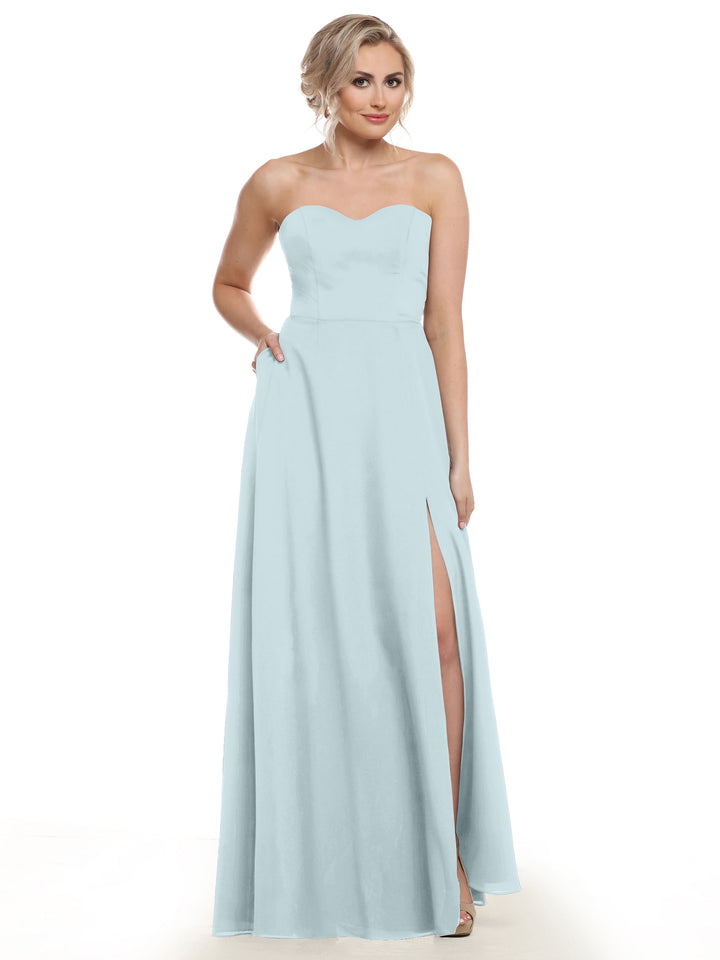 A woman models Avery Austin’s sky blue bridesmaid dress (Style 83025), featuring a strapless sweetheart neckline, floor-length a-line silhouette, thigh-high slit, and pockets—one hand in her pocket, the other at her side.