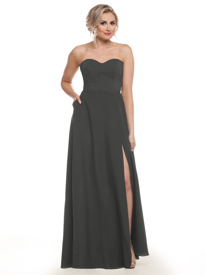 A woman models Avery Austin’s 83025: a strapless, floor-length black a-line bridesmaid dress with sweetheart neckline, high slit, and pockets, standing against a white background with her left hand in the pocket.