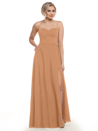A woman stands with one hand on her hip against a plain white background, modeling Avery Austin’s 83025: a floor-length, peach chiffon bridesmaid dress featuring a strapless sweetheart neckline and thigh-high slit.