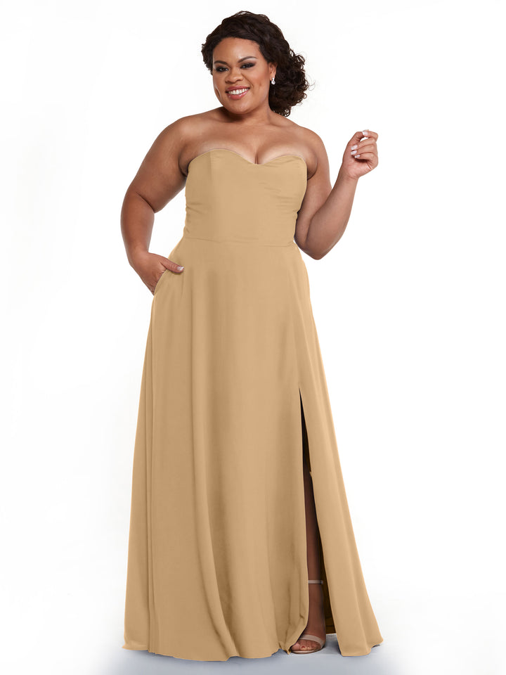 A woman wearing the Avery Austin 83025, a strapless floor-length tan chiffon bridesmaid dress with a sweetheart neckline and slit, stands smiling with one hand raised. She has curly hair and wears heels.