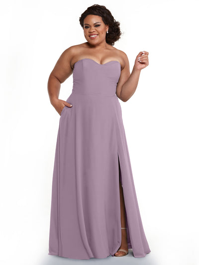 A woman models Avery Austin’s 83025: a strapless, floor-length lavender bridesmaid dress featuring a sweetheart neckline, side pockets, and thigh-high slit, standing out elegantly against a white background.