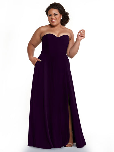 A woman smiles confidently in Avery Austin’s 83025: a deep purple, floor-length chiffon bridesmaid dress with a strapless sweetheart neckline, A-line silhouette, and thigh-high slit. She wears sandals and has elegantly styled curls.