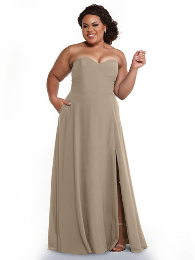 A woman poses confidently in the Avery Austin 83025: a strapless, floor-length taupe chiffon bridesmaid dress with a sweetheart neckline and side slit, smiling with one hand on her hip and the other near her face.