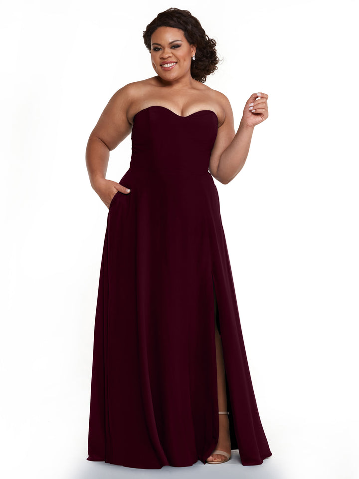 A woman smiles against a plain white background, wearing the Avery Austin 83025—a strapless burgundy bridesmaid dress with a sweetheart neckline, floor-length A-line silhouette, and thigh-high slit.