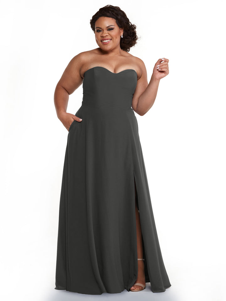 A woman in Avery Austin’s 83025—a dark gray, floor-length strapless bridesmaid dress with a sweetheart neckline and thigh-high slit—smiles, hand on hip, holding an object against a plain white background.