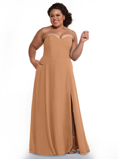 A woman smiles against a white background wearing Avery Austin’s 83025 strapless bridesmaid dress in camel, featuring a thigh-high slit and pockets. Her curly hair is styled back, one hand raised and the other resting in her pocket.