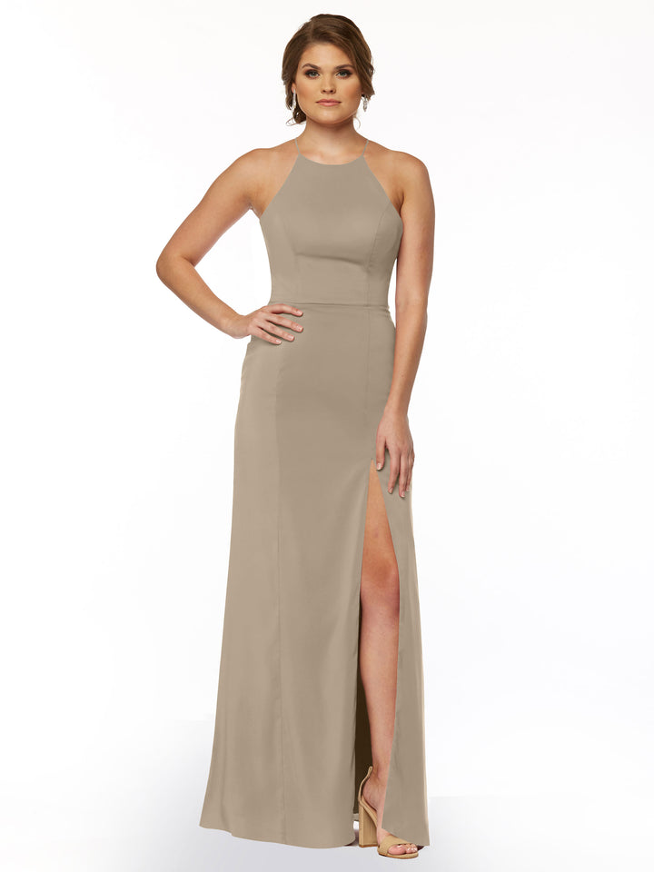 A woman with short hair models the Avery Austin 83026 dress, a sleeveless taupe sheath featuring a halter neck and front slit. She stands in high-heeled sandals, one hand on her hip and the other at her side.