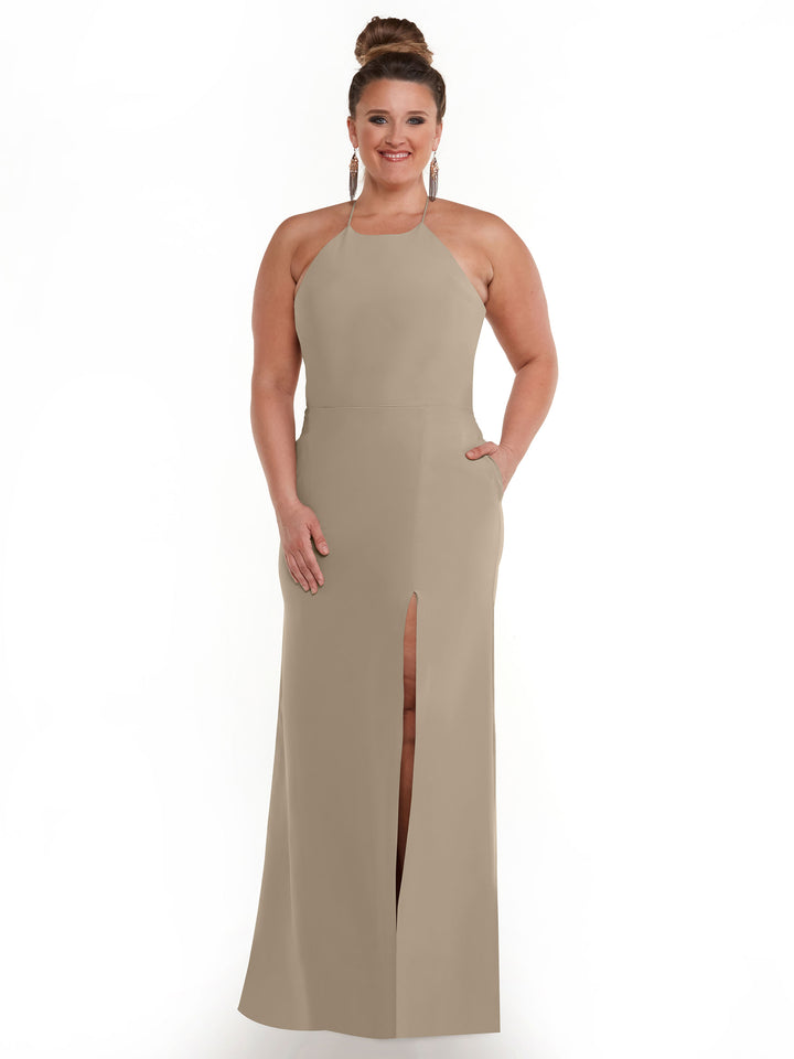 A woman smiles against a white background wearing Avery Austin’s 83026, a light beige sleeveless sheath dress featuring a halter neck, front slit, and pockets. Her hair is in a bun and she accessorizes with large hoop earrings.