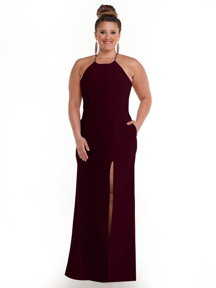 A smiling woman wears Avery Austin’s 83026, a sleeveless dark burgundy halter neck dress with pockets and a front slit, standing confidently against a white background with her hands in her pockets.