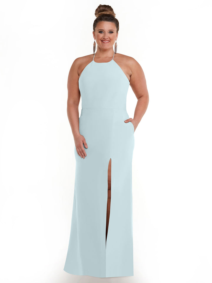 A woman smiles against a white background in Avery Austin’s 83026: a pale blue, floor-length halter neck dress with pockets and a front slit. Her hair is in a bun and her hands rest in the dress pockets.