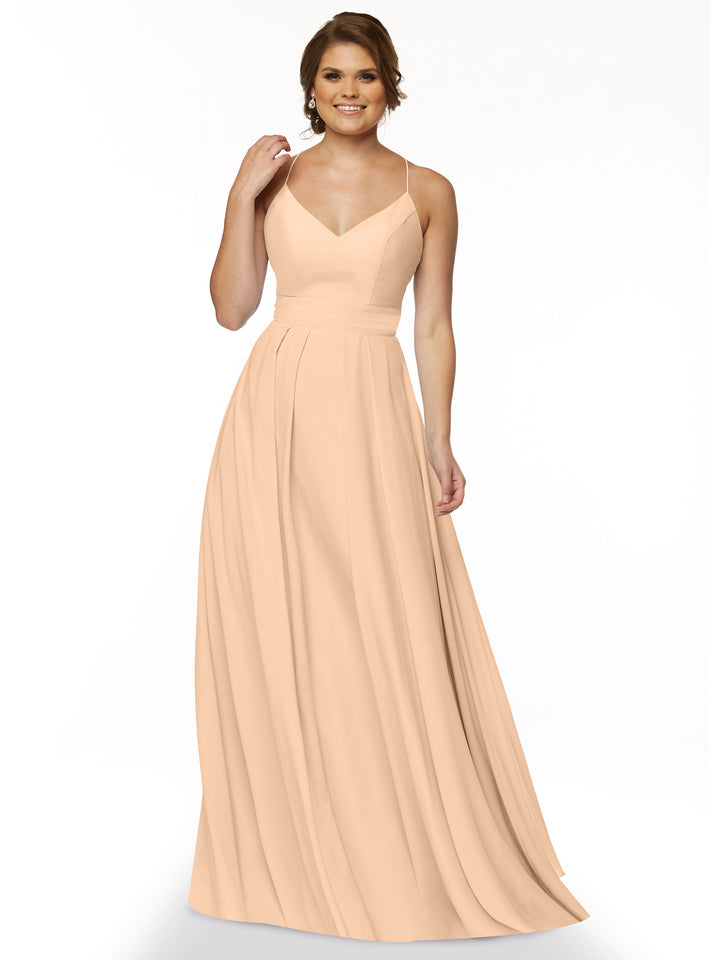 A woman stands smiling against a white background, wearing Avery Austin’s 83027: a long peach A-line dress with spaghetti straps and a fitted bodice.
