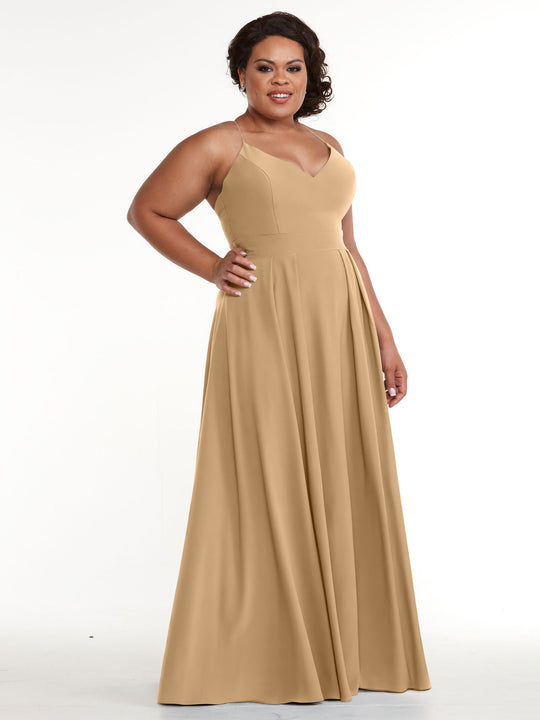 A woman stands against a plain white background, smiling confidently with one hand on her hip while wearing Avery Austin's sleeveless, floor-length tan A-Line dress (style 83027) featuring a fitted bodice and flared skirt.